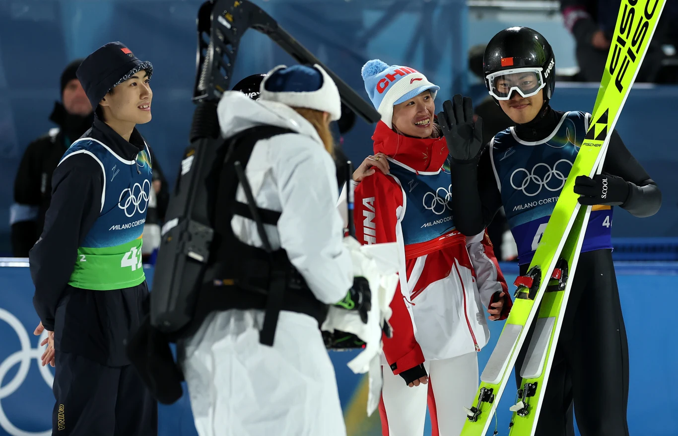 Grupa sportowców w kombinezonach i kaskach na tle charakterystycznych symboli olimpijskich, jeden z zawodników trzyma narty, wszyscy wyglądają na zadowolonych po zakończonym starcie.