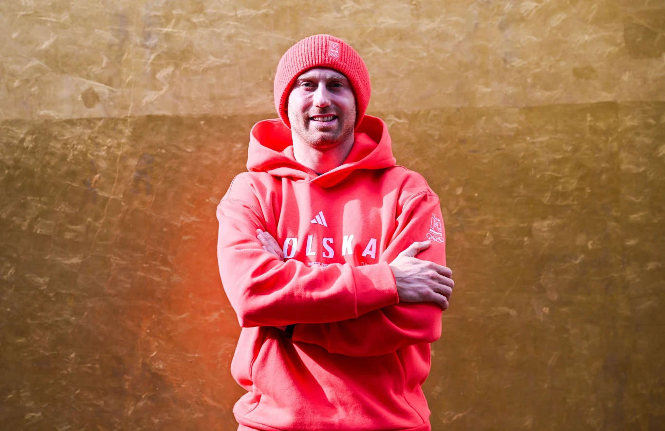 Mężczyzna w czerwonej czapce i bluzie z napisem POLSKA stoi z założonymi rękami na tle jednolitej, złocistej ściany.