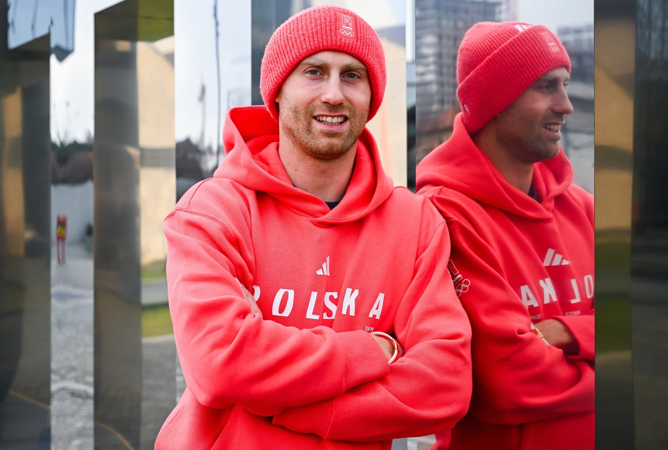 Mężczyzna w czerwonej czapce i bluzie z napisem 'Polska' stoi z założonymi rękami, uśmiechając się, obok lustra odbijającego jego sylwetkę; w tle niewyraźna zabudowa miejska.