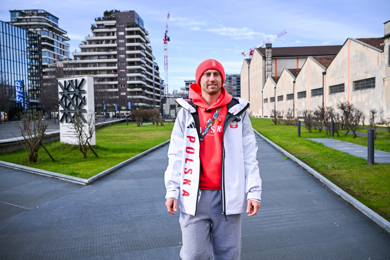 Mężczyzna ubrany w sportową kurtkę oraz bluzę i czapkę w kolorach biało-czerwonych z napisem 'Polska', stoi na tle miejskiego krajobrazu z nowoczesnymi budynkami i architekturą industrialną.