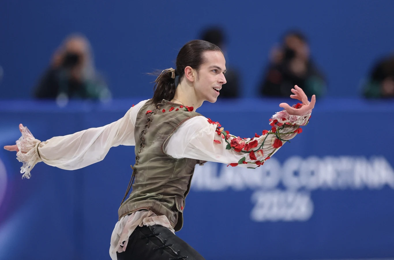 Łyżwiarz figurowy w stylizowanym kostiumie występuje na lodowisku podczas zawodów, w tle widoczny napis Milano Cortina 2026 oraz rozmazana publiczność.