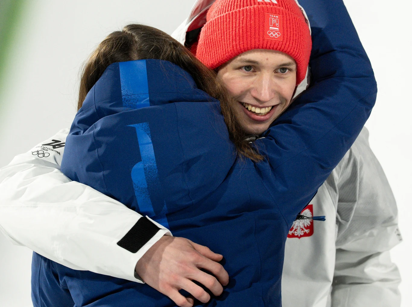 Sportowiec w czerwonej czapce i białej kurtce z orłem na piersi obejmuje osobę ubraną w niebieską kurtkę. Oboje są uśmiechnięci, atmosfera jest pełna radości i wzruszenia.