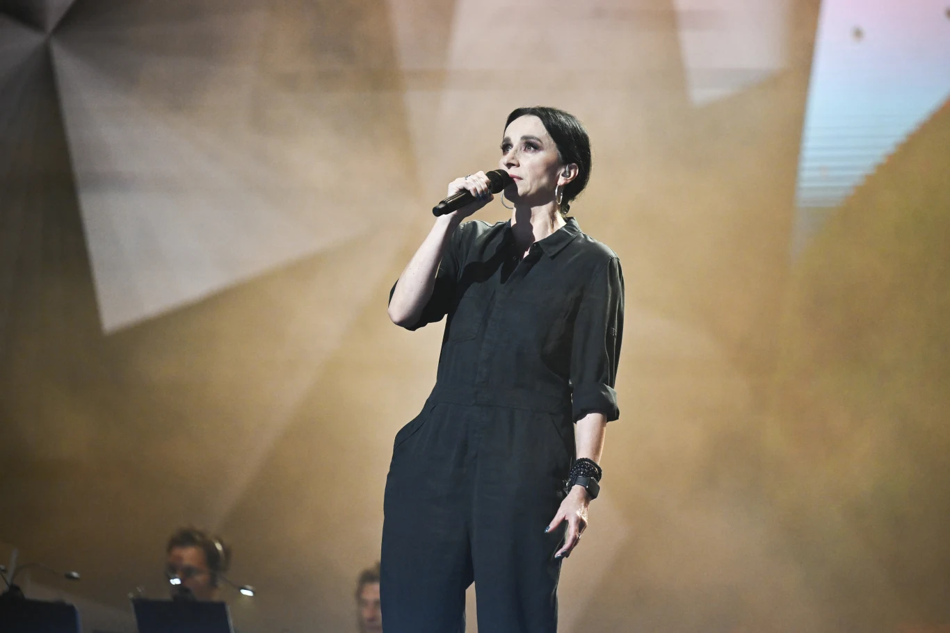 Renata Przemyk Kobieta w czarnym stroju trzyma mikrofon, występuje na scenie oświetlonej punktowym światłem, w tle widoczni są muzycy i rozmyte geometryczne kształty scenografii.