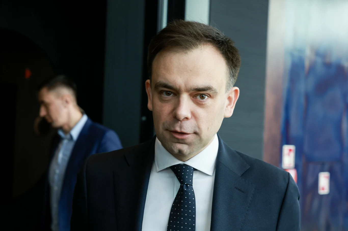 Minister finansów i gospodarki Andrzej Domański Mężczyzna w garniturze i krawacie patrzy prosto w obiektyw, w tle drugi mężczyzna w formalnym stroju odwrócony w bok, wnętrze budynku ze ścianami w ciemnych odcieniach i odbiciami światła.