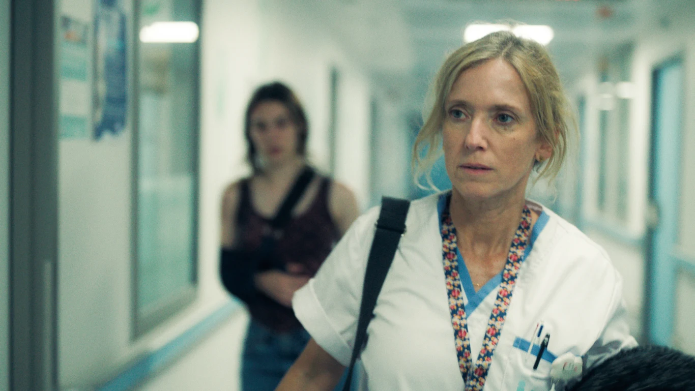 Kadr z filmu "Dla dobra Adama" Kobieta w stroju medycznym z identyfikatorem i długopisami w kieszeni idzie korytarzem szpitalnym, w tle młoda osoba z temblakiem na ramieniu.