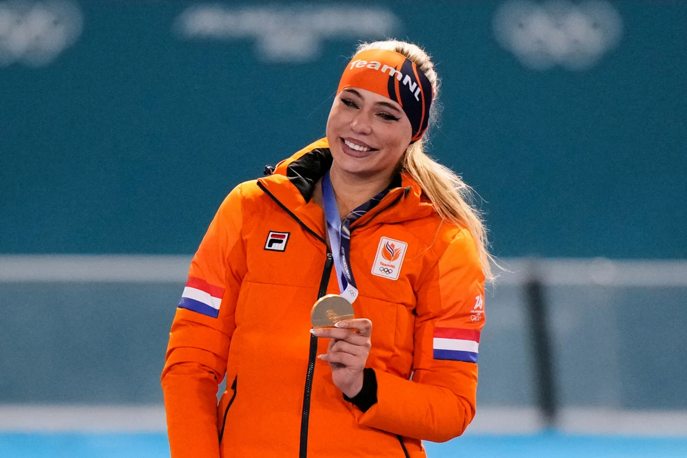 Uśmiechnięta sportsmenka ubrana w pomarańczową kurtkę i opaskę na głowie, trzymająca złoty medal na szyi. W tle rozmazane logo wydarzenia sportowego, atmosfera radości i sukcesu.