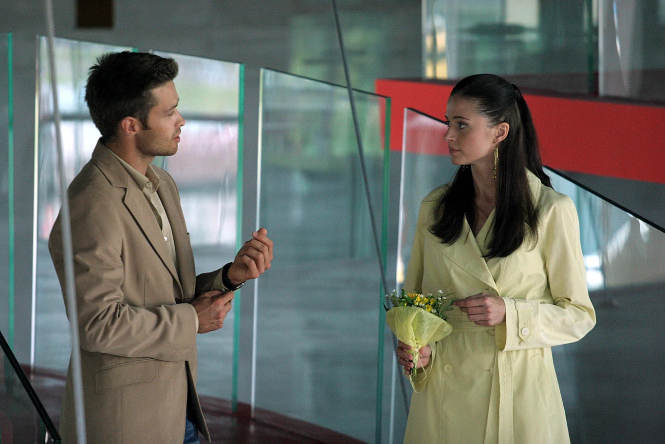 "Tylko mnie kochaj" Mężczyzna i kobieta prowadzą rozmowę w nowoczesnym, przeszklonym wnętrzu, kobieta trzyma w rękach niewielki bukiet kwiatów.