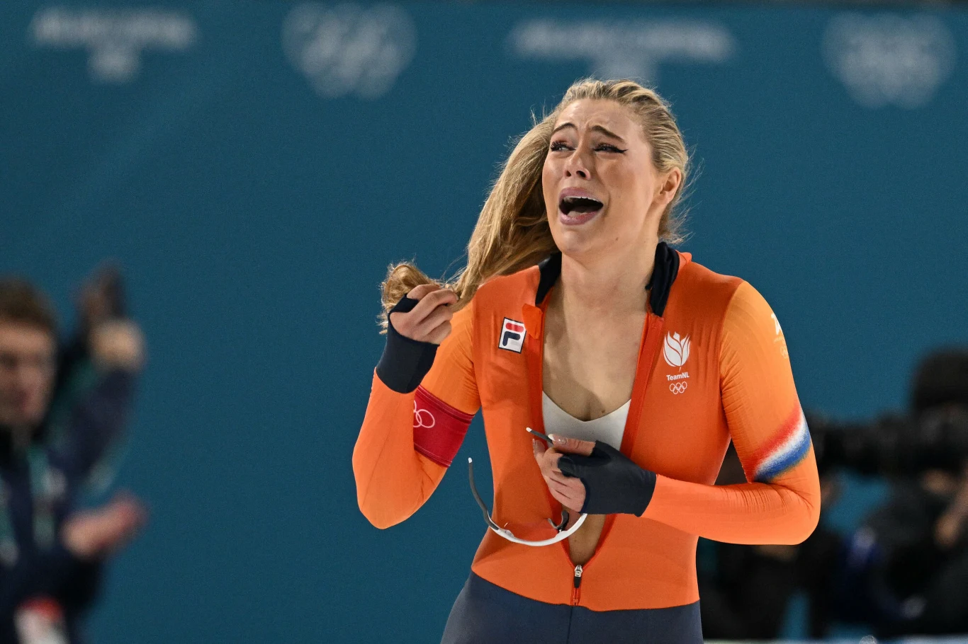 Zawodniczka ubrana w pomarańczowy strój sportowy, z wyrazem silnych emocji na twarzy, znajduje się na arenie sportowej, trzymając medal lub identyfikator.