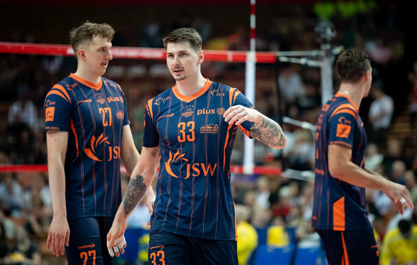 Zawodnicy siatkarskiej drużyny JSW ubrani w klubowe granatowe stroje z pomarańczowymi elementami, stojący blisko siebie na boisku w trakcie meczu, w tle widoczne siatka oraz rozmazane postacie kibiców.