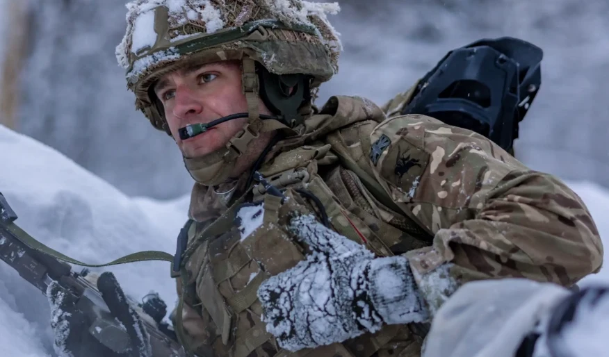 Wielka Brytania przerzuci do Norwegii dodatkowy tysiąc żołnierzy (zdj. ilustracyjne) Żołnierz w kamuflażu zimowym z twarzą częściowo pokrytą śniegiem, trzymający broń i przygotowany do działania w zaśnieżonym terenie.