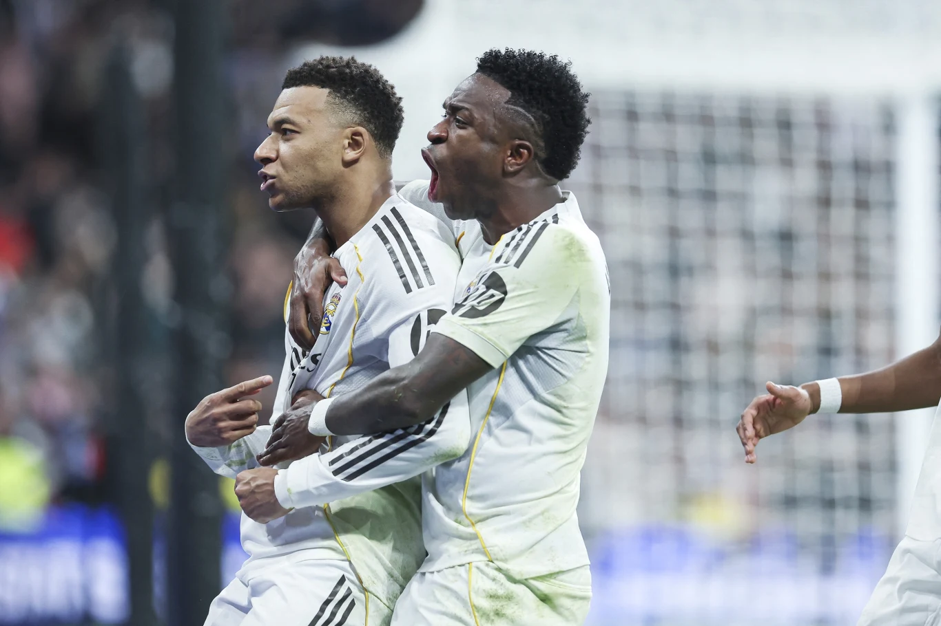 Dwaj piłkarze w białych strojach drużynowych świętują zdobycie bramki podczas meczu, jeden z nich obejmuje swojego kolegę z zespołu, okazując silne emocje.