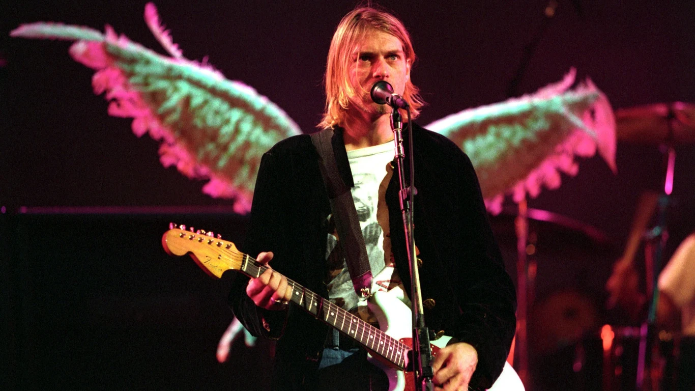 Kurt Cobain (Nirvana) Mężczyzna z gitarą elektryczną stoi na scenie, w tle widoczne duże dekoracyjne skrzydła, które sprawiają wrażenie, jakby wyrastały z jego pleców. Scena oświetlona kolorowymi światłami.