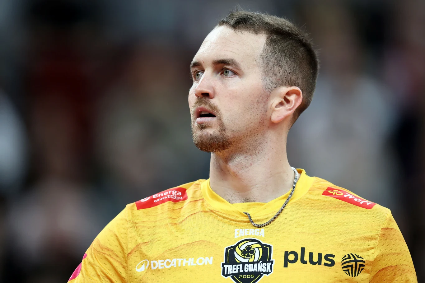 Młody mężczyzna w żółtej sportowej koszulce z logo klubu Trefl Gdańsk, patrzący w bok, na szyi srebrny łańcuszek, tło rozmyte.