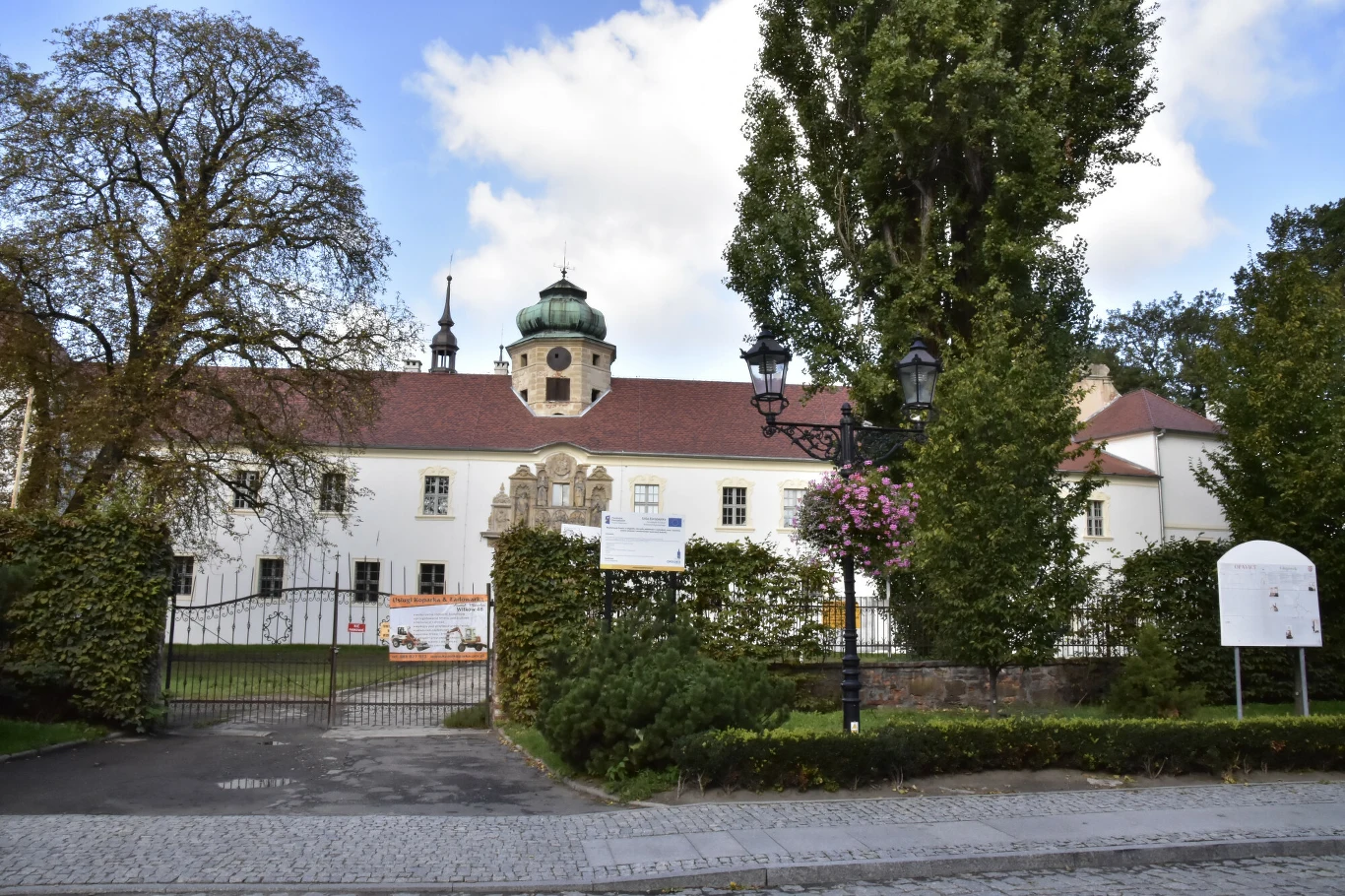 Zamek w Głogówku Zabytkowy pałac z białą elewacją i czerwonym dachem otoczony zielenią, bramą wjazdową oraz stylowymi latarniami, na tle błękitnego nieba z chmurami.