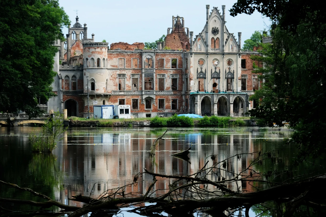 Pałac w Kopicach Zabytkowy, częściowo zrujnowany pałac z cegły odbijający się w spokojnej tafli wody, otoczony zielenią i drzewami, z widocznymi śladami zniszczeń oraz pozostałościami dawnych elementów architektonicznych.