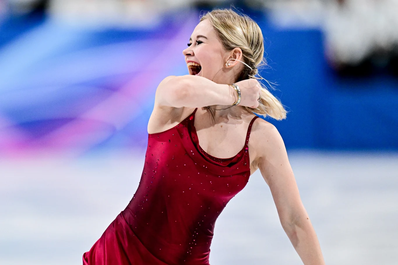 Łyżwiarka figurowa ubrana w bordową suknię wykonuje efektowny gest z uśmiechem, stojąc na lodowisku podczas zawodów sportowych.