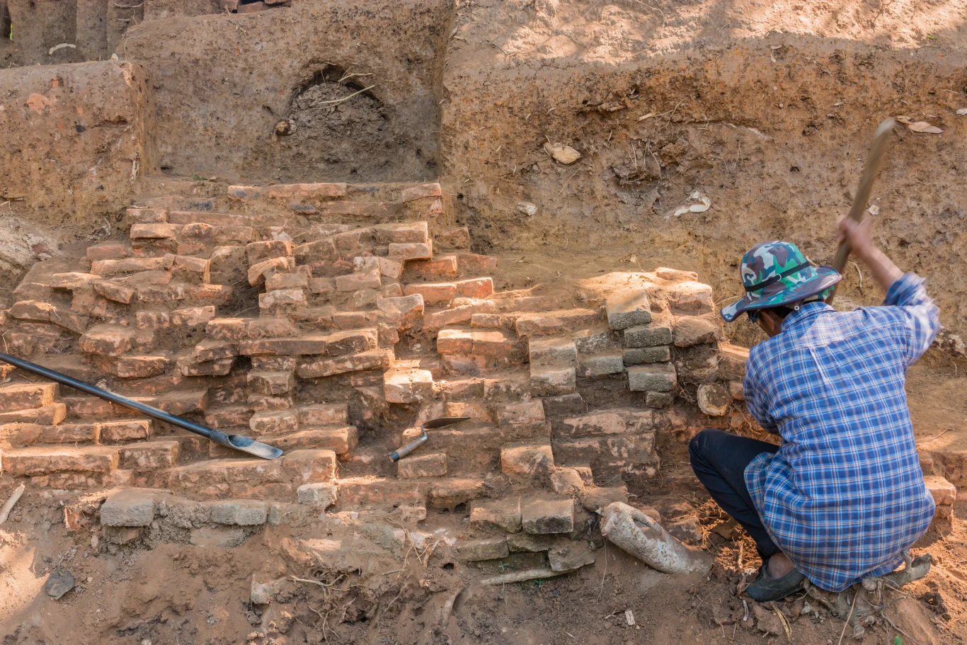 Nowe odkrycie w stolicy Kambodży. Zdj. pogl. Osoba w kapeluszu przy pracy w wykopie archeologicznym, odsłania ceglane fundamenty w Phnom Penh.