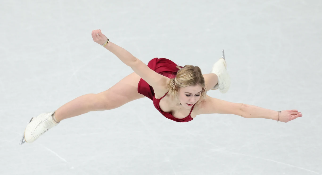 Łyżwiarka figurowa w czerwonym stroju wykonująca dynamiczny skok z rozłożonymi ramionami i nogami na lodowisku.