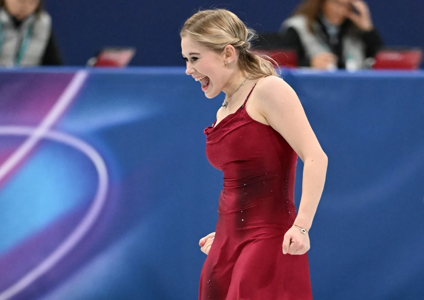 Łyżwiarka figurowa w czerwonej sukni wyraża radość podczas występu na lodowisku, w tle rozmyty niebiesko-fioletowy baner i publiczność.