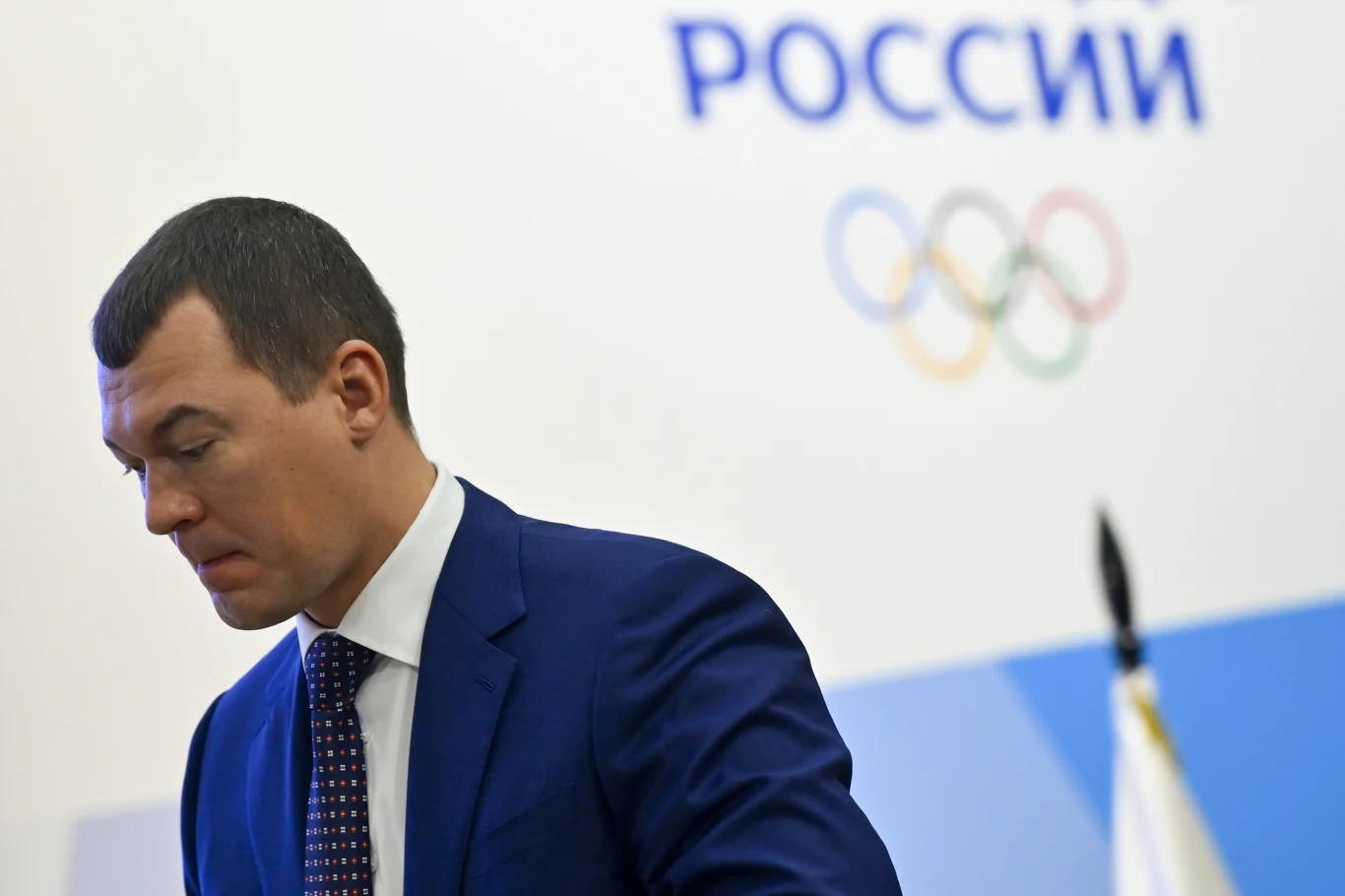 Mężczyzna w garniturze stoi na tle białej ściany z napisem „Rosja” oraz symbolami olimpijskimi; po prawej stronie widoczny fragment flagi.