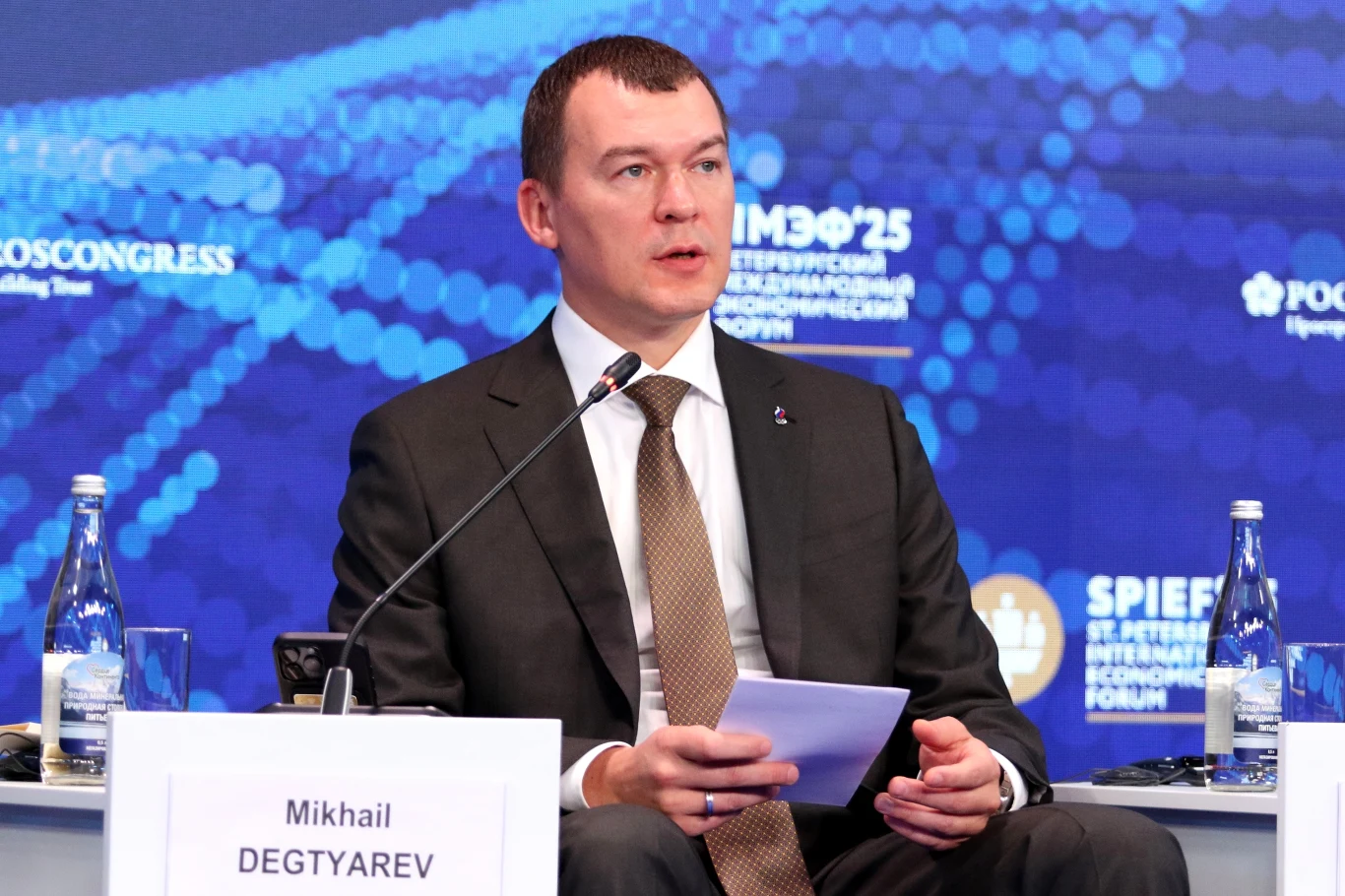 Mężczyzna w garniturze siedzi przy stole z mikrofonem podczas konferencji, trzyma kartkę papieru, przed nim stoi tabliczka z imieniem i nazwiskiem. W tle niebieska ścianka z logo wydarzenia i kilka butelek wody na stole.