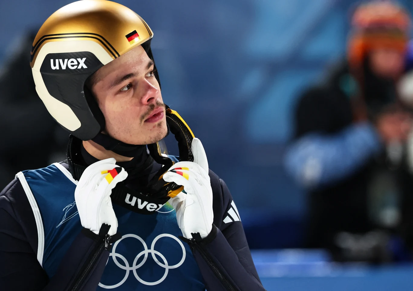 Sportowiec w hełmie i kombinezonie z niemiecką flagą oraz olimpijskimi pierścieniami przygotowuje się do startu, skoncentrowany, trzyma pasek kasku i patrzy w bok.