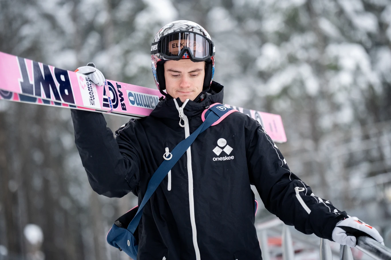 Młody mężczyzna w czarnej kurtce i kasku narciarskim trzyma różowe narty na ramieniu, idąc przez zaśnieżony teren, w tle widoczne rozmyte drzewa.