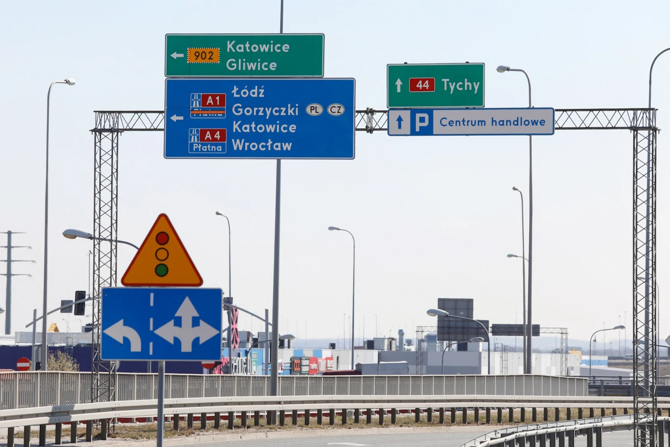 Zestaw znaków drogowych i tablic kierunkowych wskazujący różne kierunki jazdy oraz informacje o autostradach, centrum handlowym i pobliskich miastach, takich jak Katowice, Gliwice, Tychy i Łódź. Widoczne fragmenty jezdni, barierki drogowe oraz infrastr...