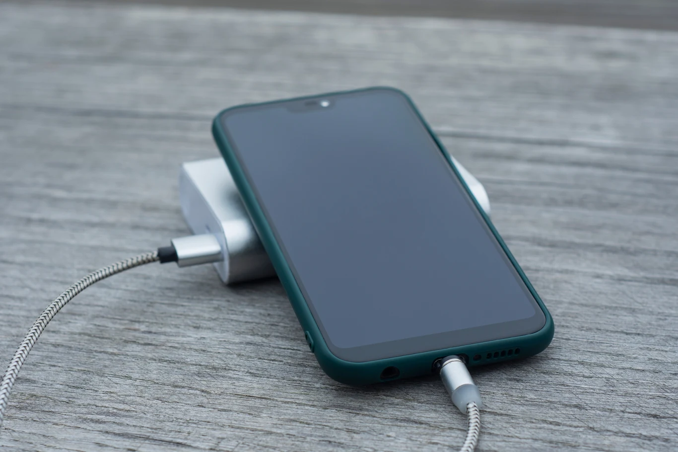 Smartfon w ciemnozielonym etui podłączony do powerbanku i kabla, leżący na drewnianej powierzchni.