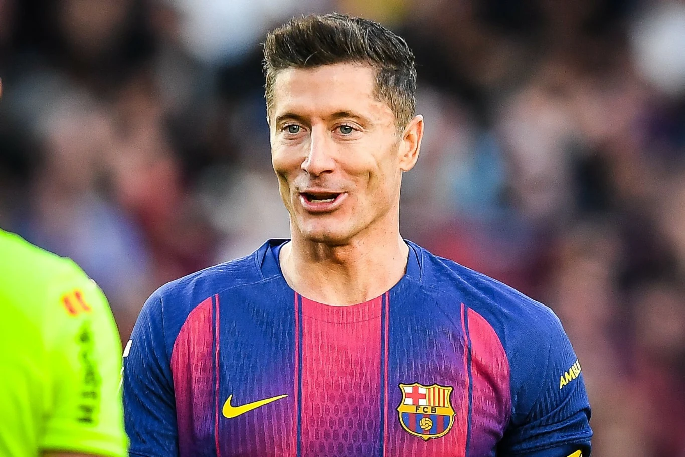 Robert Lewandowski Piłkarz w koszulce FC Barcelony z numerem na rękawie podczas meczu, uśmiechnięty, w tle rozmyte postacie i trybuny stadionu.