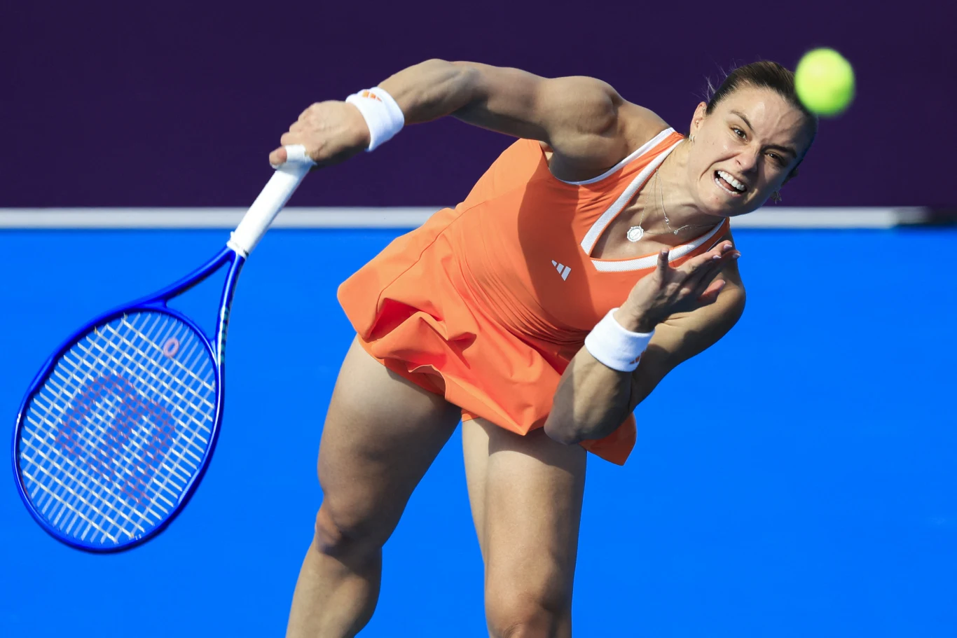 Maria Sakkari, Doha 2026 Tenisistka w pomarańczowym stroju wykonuje serwis podczas meczu, skupiona, dynamiczny ruch, niebieski kort w tle, lotka tenisowa unosząca się w powietrzu.