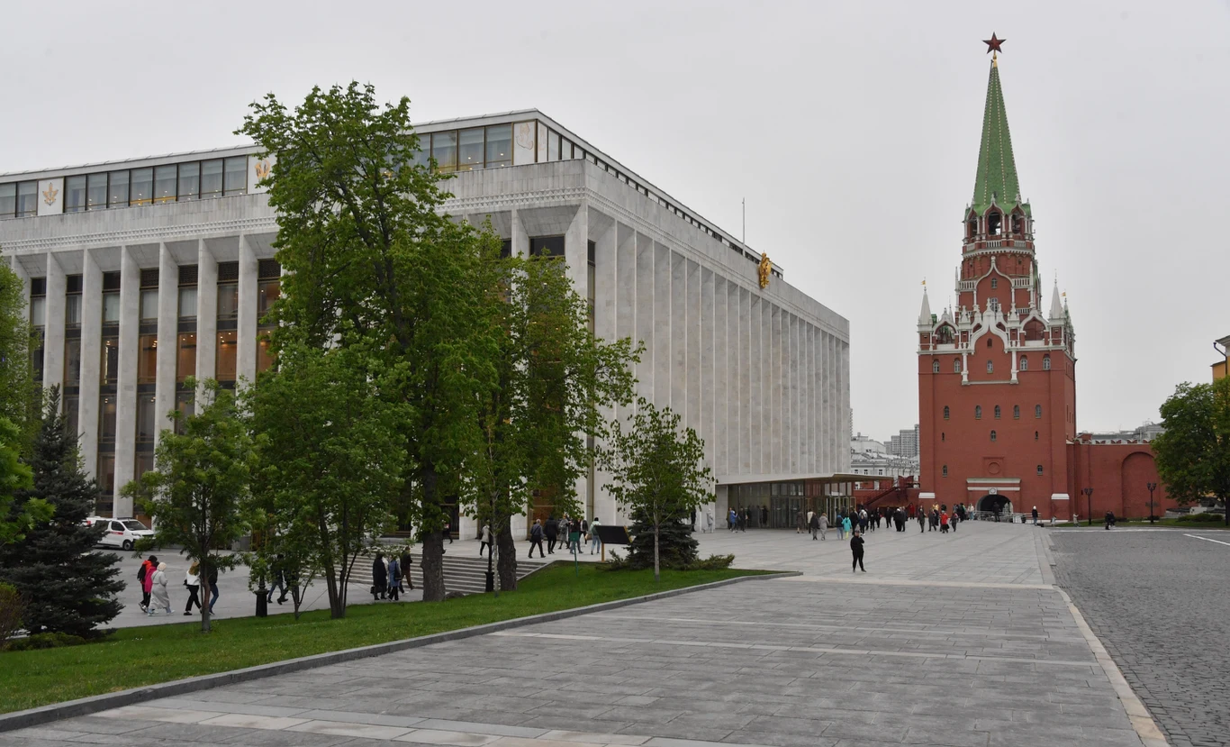 Plac z nowoczesnym budynkiem po lewej stronie, otoczony zielenią, z charakterystyczną czerwoną wieżą z zielonym dachem i czerwoną gwiazdą po prawej stronie, po placu spacerują ludzie.