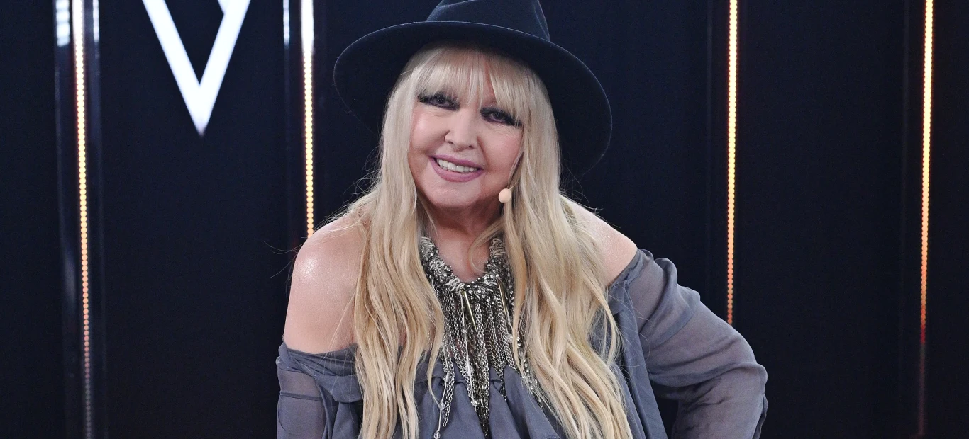 Maryla Rodowicz Kobieta z długimi blond włosami uśmiecha się, ubrana jest w luźną szarą bluzkę odsłaniającą ramiona, duży naszyjnik i czarny kapelusz. W tle widoczne są pionowe linie świetlne i geometryczny wzór.