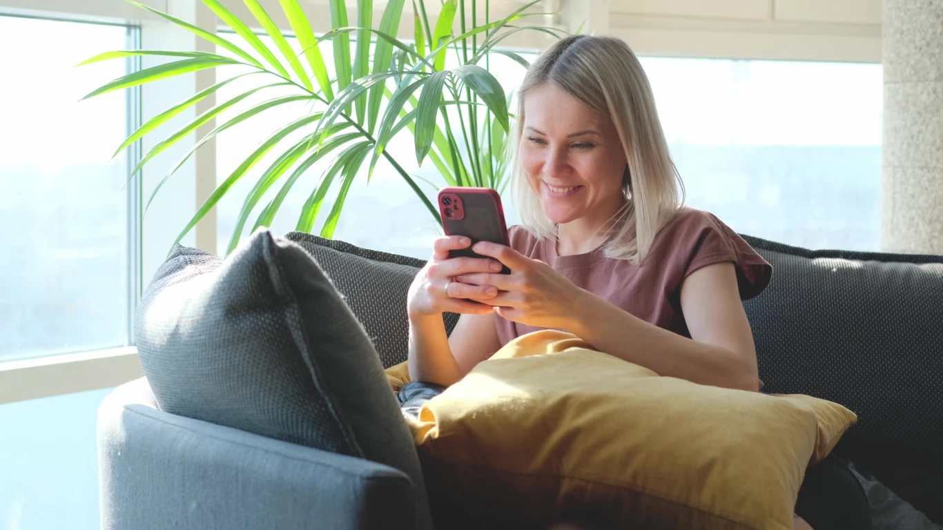 Uśmiechnięta kobieta siedzi wygodnie na kanapie z jasną poduszką w dłoniach, trzymając smartfon i patrząc na ekran. W tle widoczne są duże okna oraz zielona roślina.