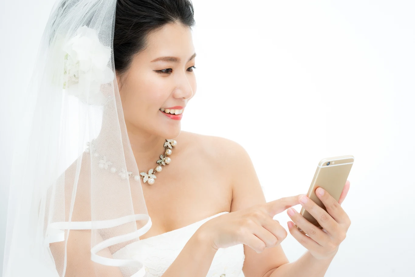 Uśmiechnięta panna młoda w sukni ślubnej i welonie, ozdobiona kwiatową biżuterią, korzysta z telefonu komórkowego na jasnym tle.