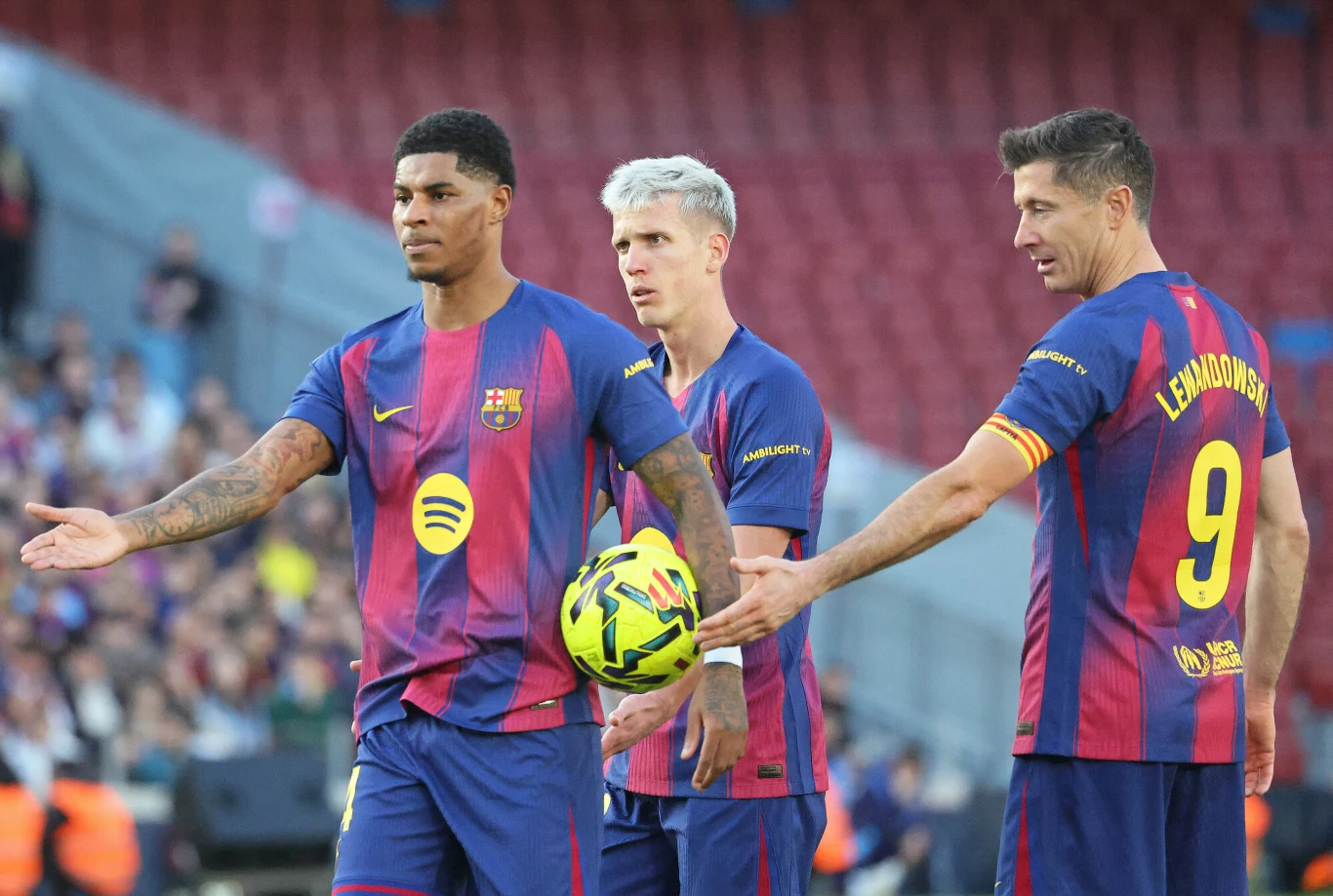 Trzech piłkarzy w koszulkach FC Barcelona na boisku piłkarskim, jeden z nich trzyma piłkę, a widownia w tle obserwuje wydarzenie.