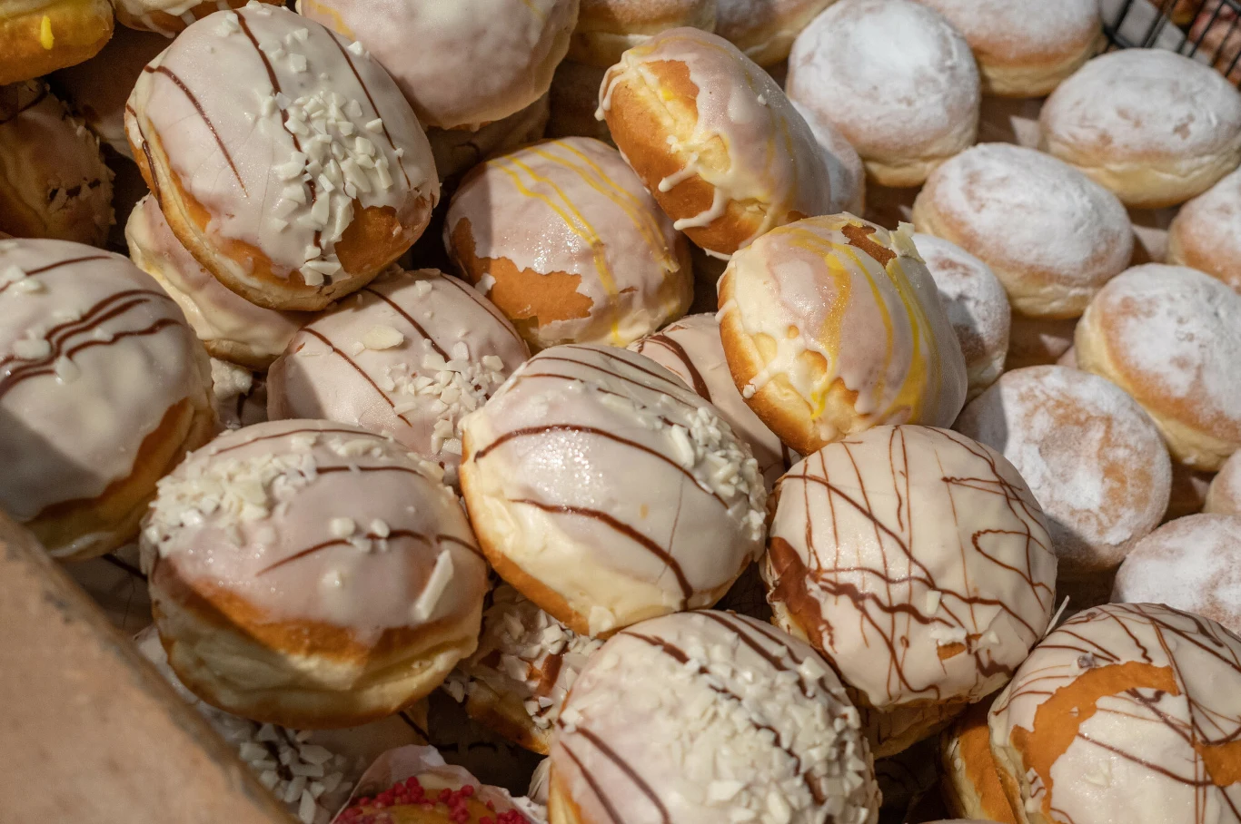 Stos pączków z różnymi polewami i posypkami, niektóre dekorowane białą czekoladą i polewą czekoladową, inne oprószone cukrem pudrem.