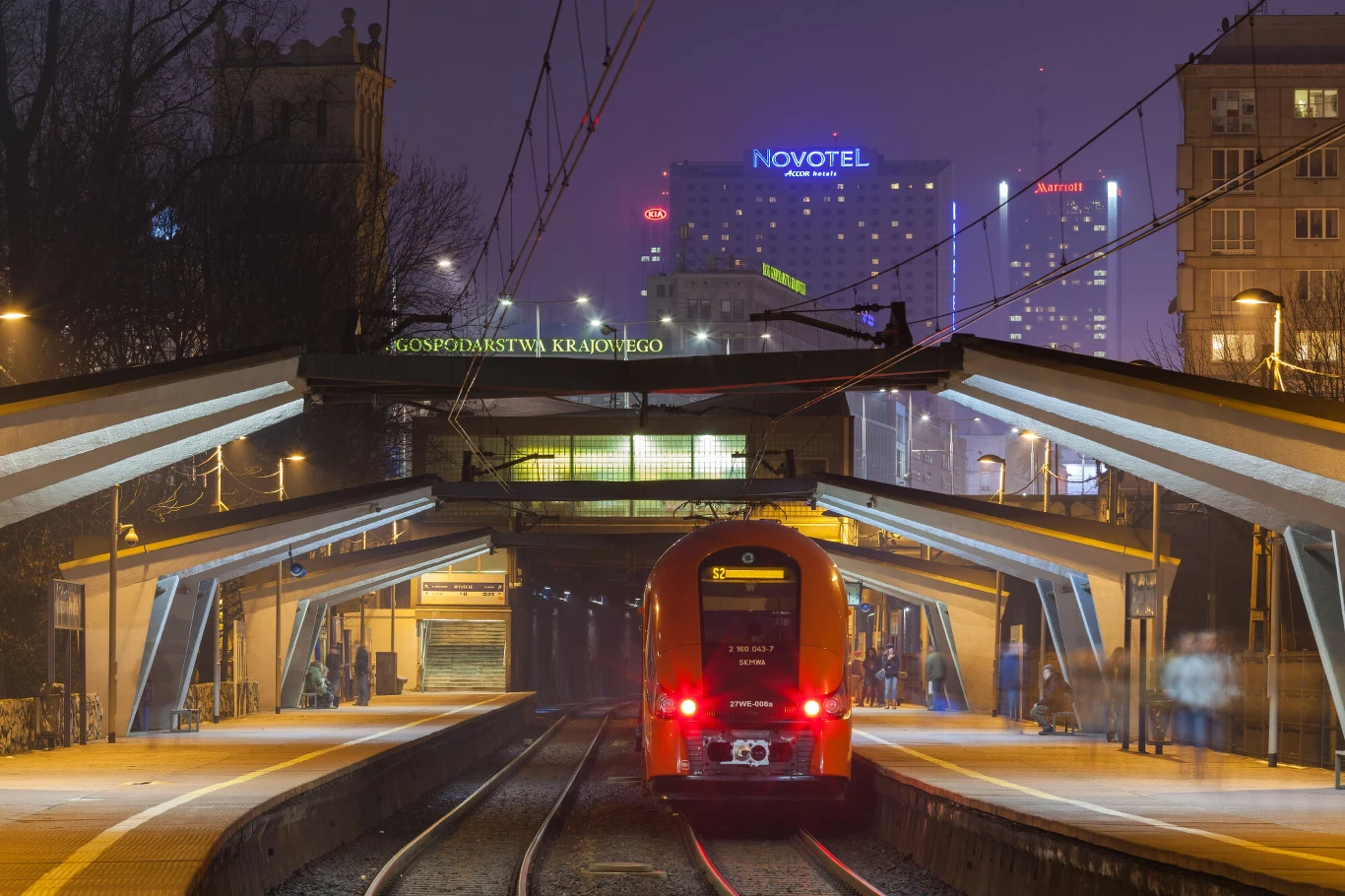 Wjazd do tunelu linii średnicowej przy przystanku Warszawa Powiśle Pociąg osobowy stoi na peronie stacji Warszawa Ochota przy miejskich światłach nocą, w tle widoczne są oświetlone budynki z wyraźnym napisem Novotel na jednym z nich.
