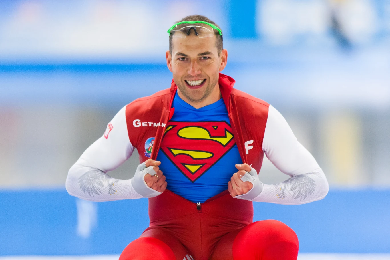 Sportowiec w stroju z barwami narodowymi, odsłaniający pod kombinezonem koszulkę z logo Supermana, uśmiechnięty, na tle lodowiska.
