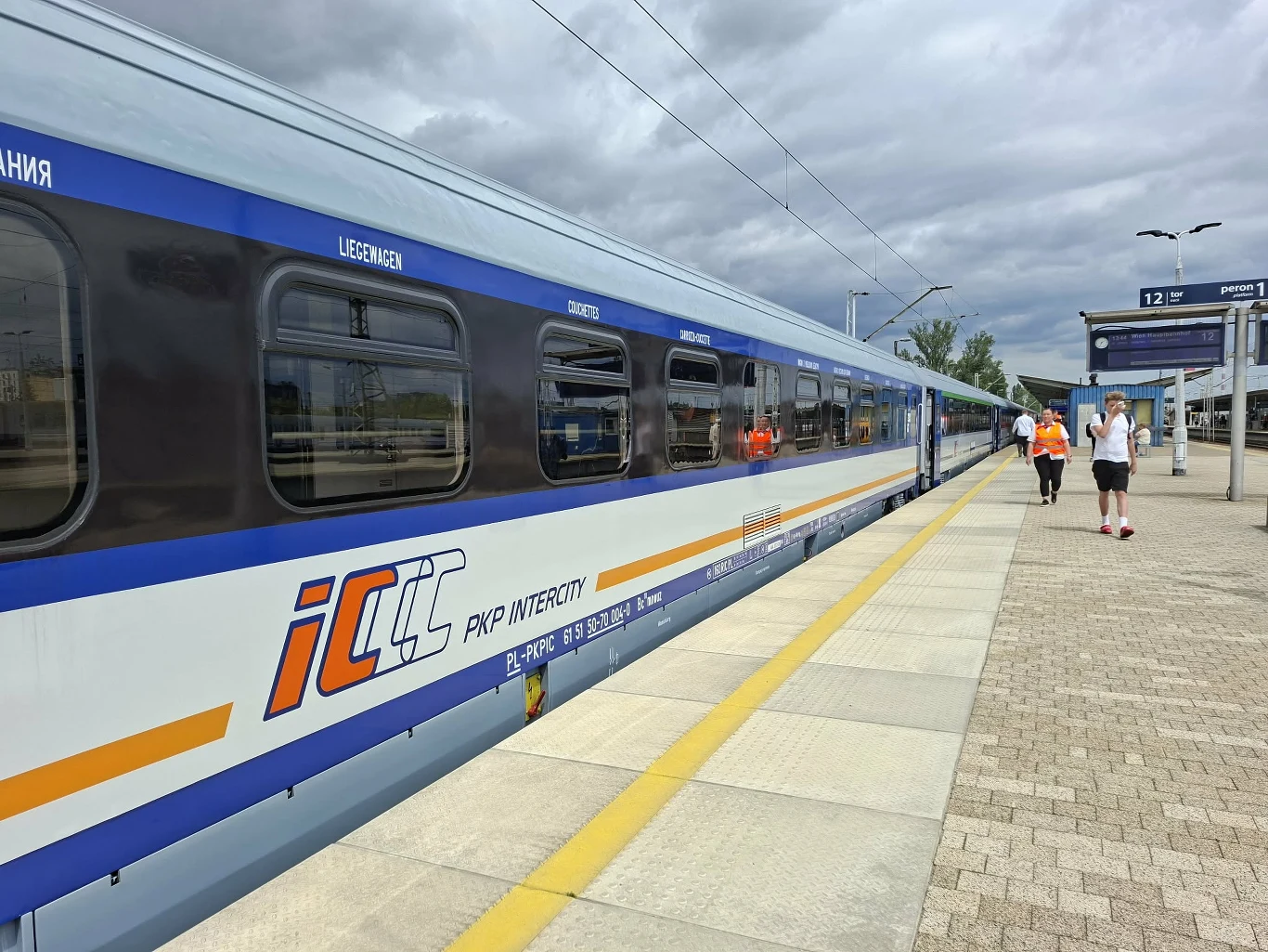 Wagon pociągu PKP Intercity stojący przy peronie stacji, na peronie kilka osób wchodzących lub wychodzących z pociągu, częściowo pochmurne niebo.