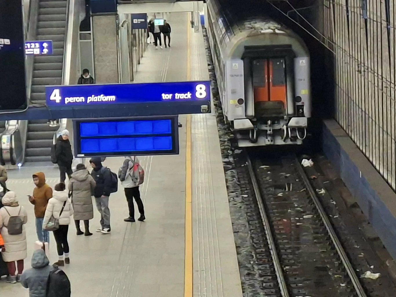 Peron kolejowy z grupą oczekujących pasażerów, widoczny pociąg stojący przy torze oraz elektroniczne tablice informacyjne wskazujące numer peronu i toru. Atmosfera codziennego ruchu na dworcu, ludzie ubrani w zimowe ubrania.