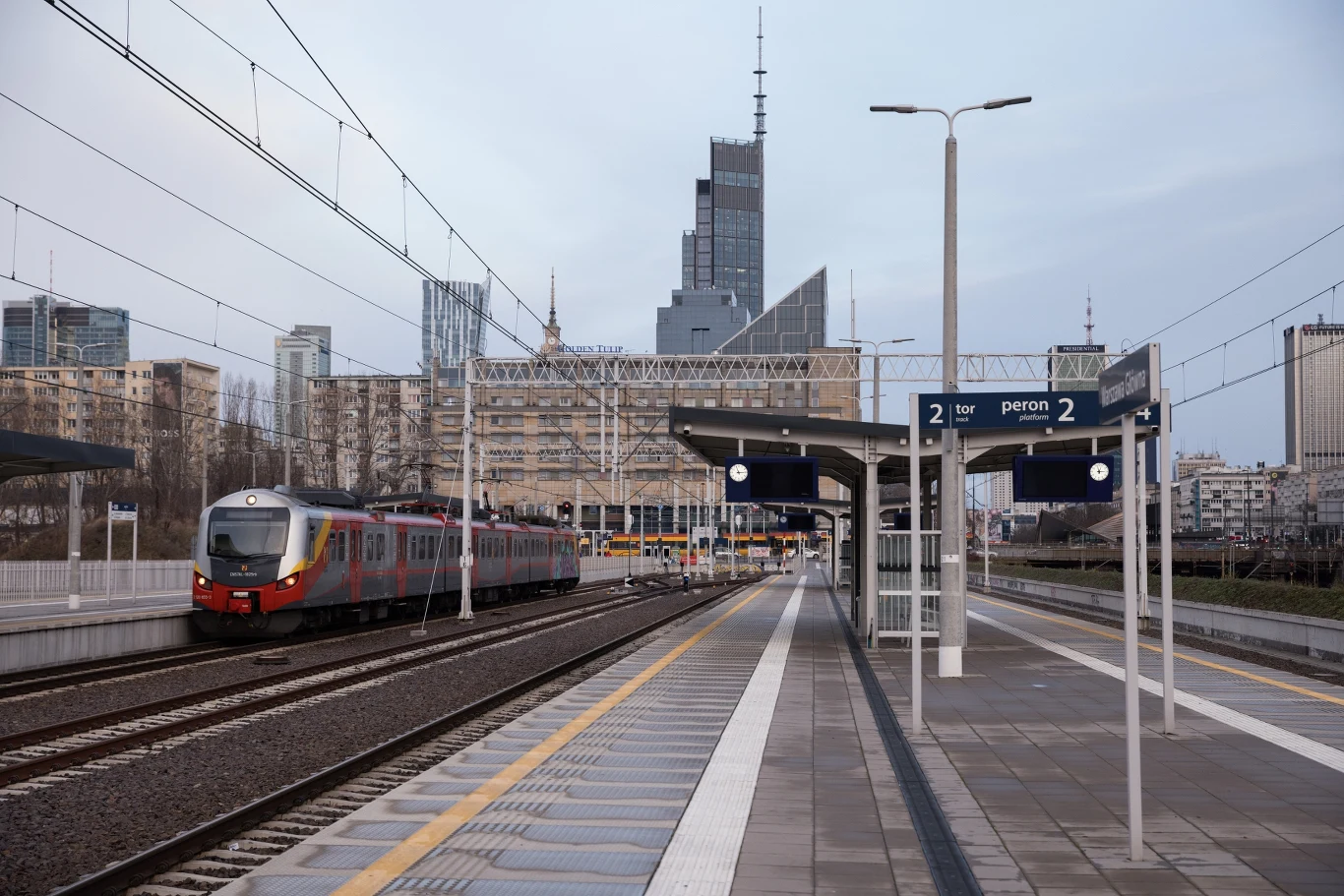 Nowoczesny pociąg przy peronie miejskiej stacji na tle wysokich budynków i wieżowców, otoczony infrastrukturą kolejową i elementami miejskiej architektury.