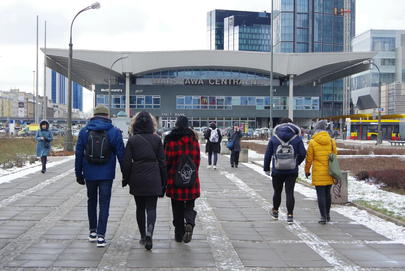 Warszawa Centralna to kluczowe miejsce w stolicy. W ciągu nadchodzących lat dworzec ma zmienić swoje oblicze Ludzie ubrani w zimowe kurtki idą pieszo chodnikiem w kierunku wejścia do Dworca Centralnego w Warszawie, w tle nowoczesne wieżowce i minimalne ilości śniegu na ziemi.