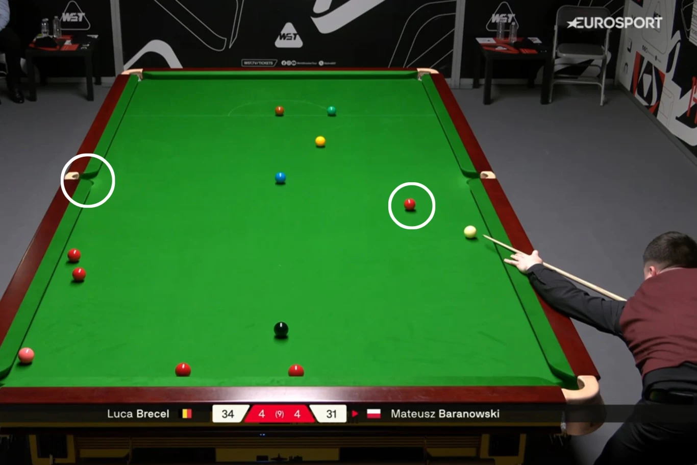 Rozgrywka snookera z widocznym stołem, kilkoma bilami oraz zawodnikiem przygotowującym się do uderzenia. Na ekranie widać aktualny wynik meczu oraz nazwiska graczy. Obraz uchwycony jest z góry, pokazując całą przestrzeń stołu bilardowego.