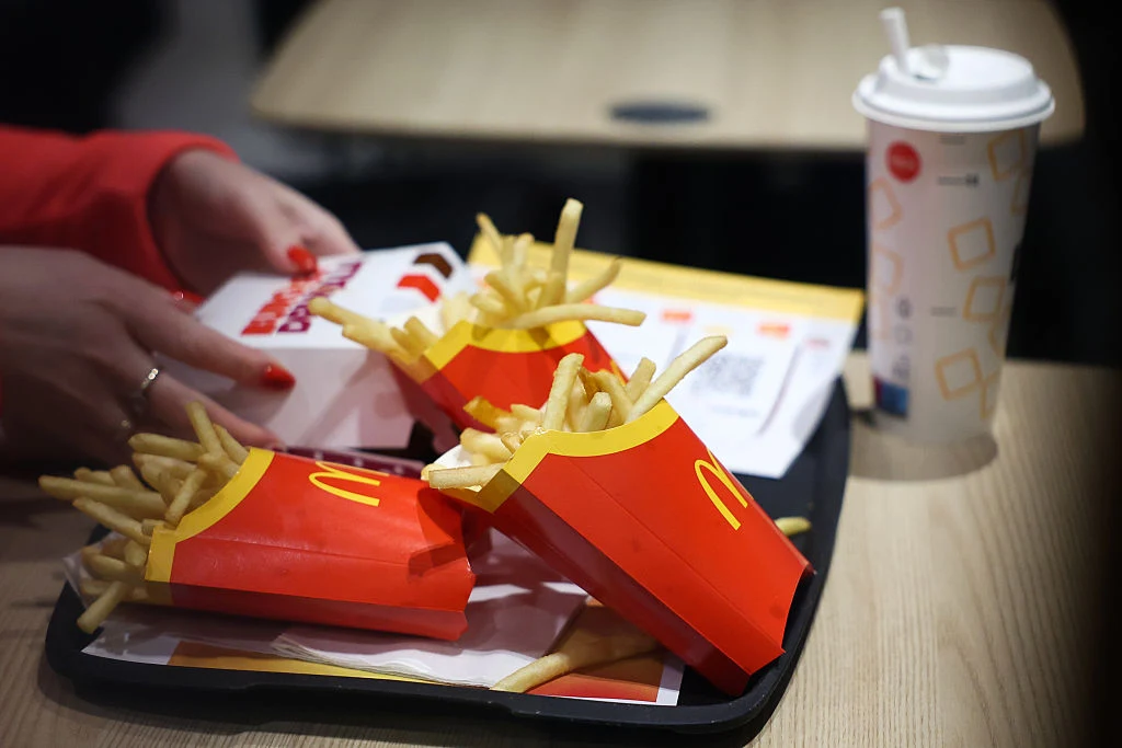Jedzenie z McDonald's Taca z trzema porcjami frytek w czerwonych opakowaniach z logo McDonald's, napojem w papierowym kubku ze słomką oraz dłonią osoby sięgającą po pudełko z jedzeniem. Wszystko znajduje się na stole w lokalu gastronomicznym.