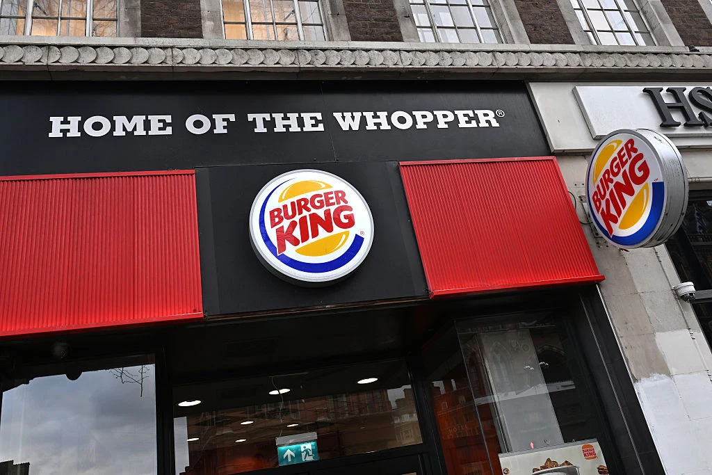Restauracja Burger King Widoczna fasada restauracji Burger King z charakterystycznym logo oraz napisem HOME OF THE WHOPPER na tle czarnej tablicy, otoczona elementami budynku.