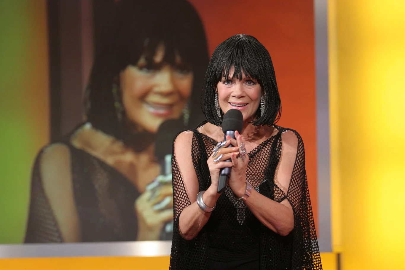 Izabela Skrybant-Dziewiątkowska Kobieta z ciemnymi włosami trzyma mikrofon, uśmiecha się i stoi na tle dużego ekranu, na którym wyświetlany jest jej portret w zbliżeniu. Tło w odcieniach żółci i pomarańczu, a jej strój to czarna kreacja z odkrytymi ramionami.