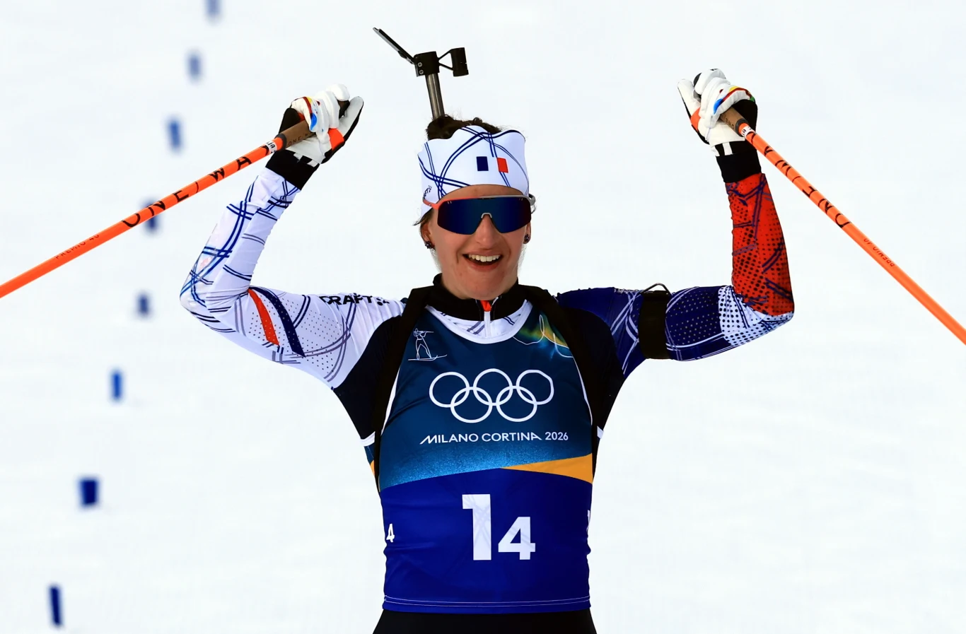 Julia Simon Biathlonista w stroju sportowym i okularach, z numerem 14 na piersi oraz z karabinem na plecach, unosi kijki w geście triumfu po ukończeniu wyścigu podczas zimowych igrzysk olimpijskich.