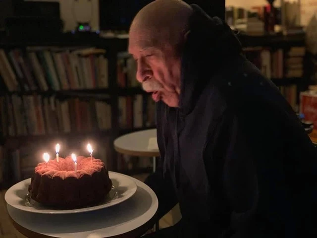 Starszy mężczyzna z pochyloną głową zdmuchuje świeczki na czekoladowym torcie, wokół półki z książkami i nastrojowe oświetlenie.