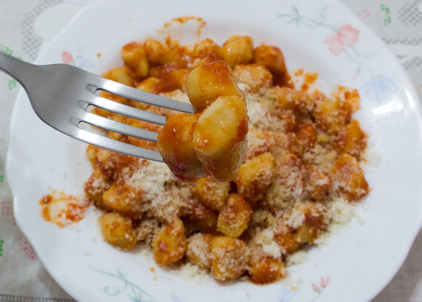 Gotowe do ugotowania gnocchi możesz kupić w większości sklepów Kluski gnocchi polane sosem pomidorowym, posypane tartym serem, na białym talerzu z kwiatowym wzorem; na pierwszym planie widelec unoszący porcję kluski.
